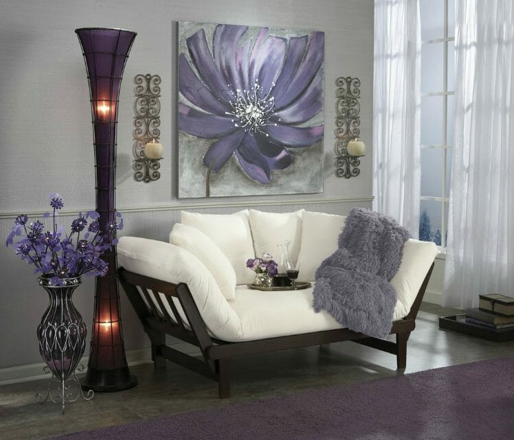 Room setting in white and shades of purple, with a futon, large floral canvas, lit floor lamp, shaggy throw, and flowers.