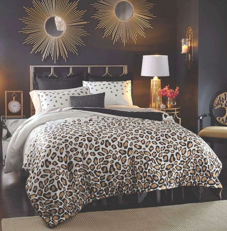 A bedroom setting in white, black and gold, with an animal print bed set, two starburst mirrors, and a lit lamp on a table.