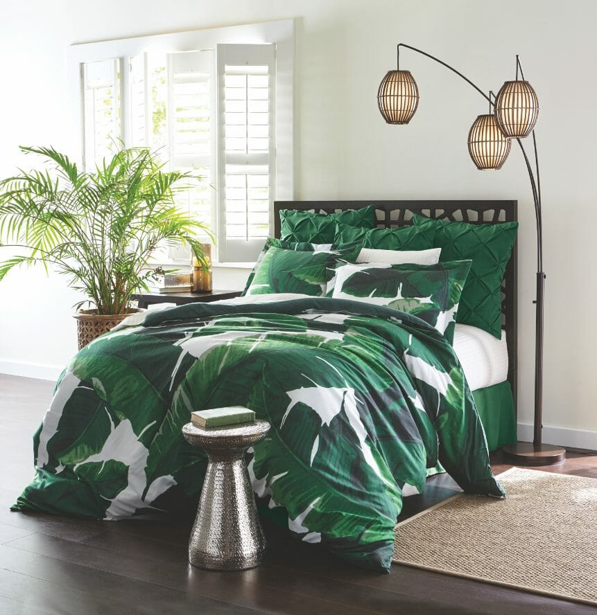An emerald green and white bed set of palm fronds, a floor lamp with three globes, a potted palm, and a silver table.