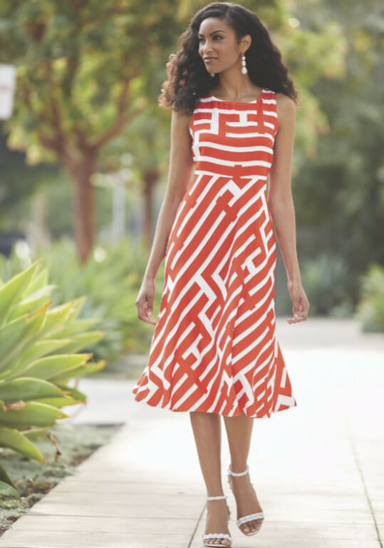 A long-haired black woman in an orange and white graphic print A-line dress, white sandals, and white drop earrings.