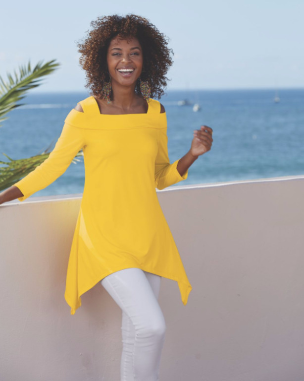 A laughing seaside black woman wearing a bright yellow cold shoulder tunic, white skinny jeans, and long drop earrings.