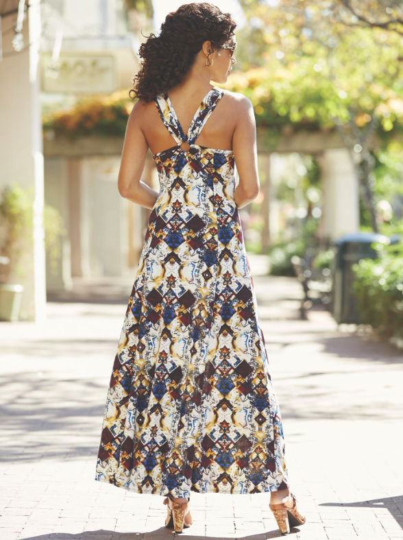 A back view of a woman with long curly hair, wearing a halter style long dress in a white, navy, brown and tan print.