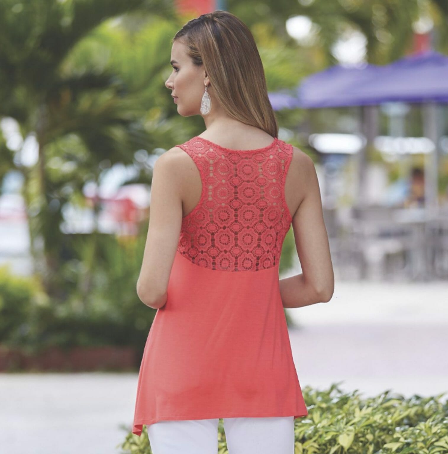 Back view of a woman outdoors, wearing a coral lace back sleeveless top, white drop earrings, and white pant.