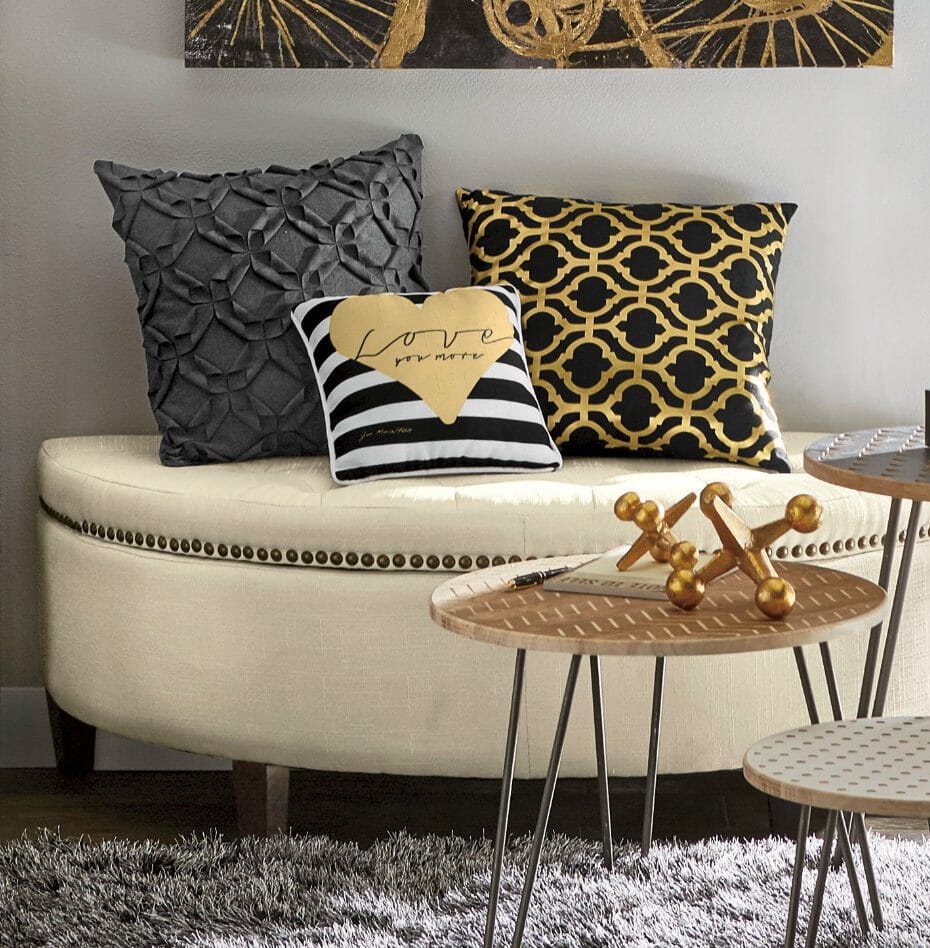 Three different decorative pillows on a white storage bench, in black, white, and gold, and a table with large gold jacks.