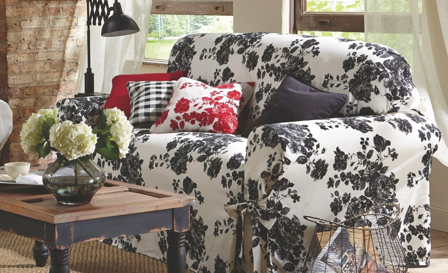 A sofa slipcover in a white and black floral print, red toss pillows, and a coffee table with vased white hydrangeas. 