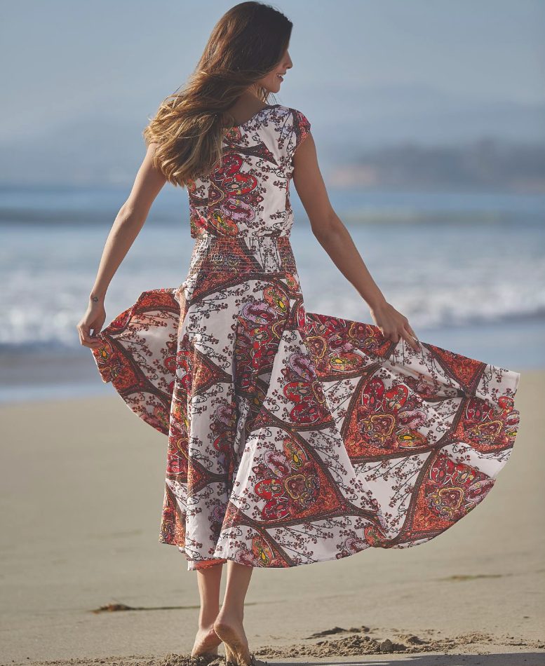 Back view of a woman on a windy ocean beach, wearing a full-skirted summer dress in a white, orange, red and purple pattern.