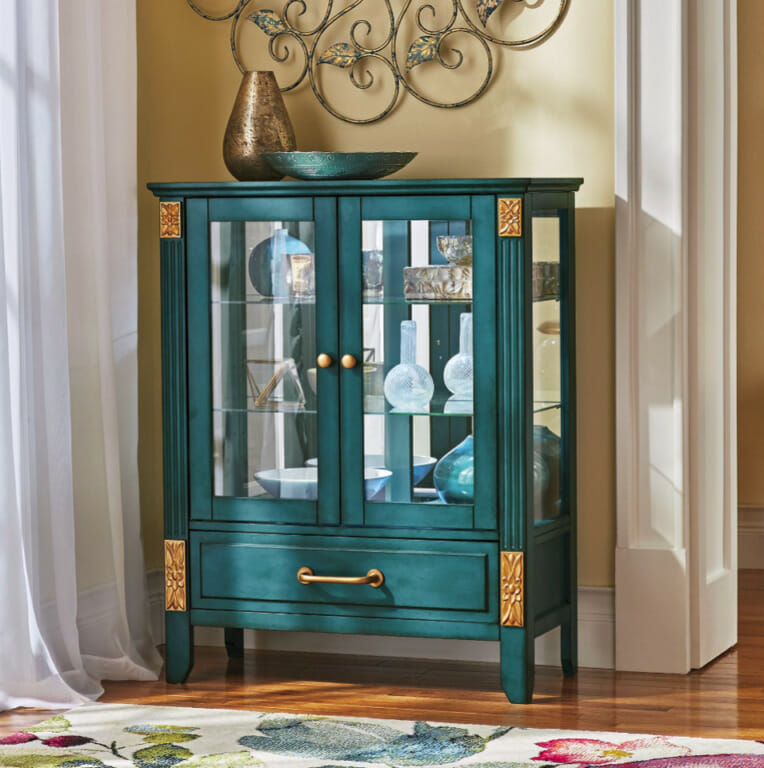 A filled turquoise etagere with a mirror back, a drawer and two glass doors, gold handles and accents, and art bowls on top.