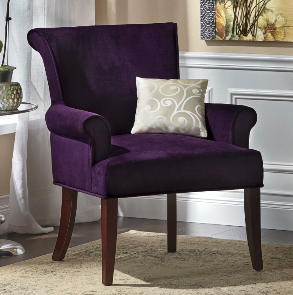 A purple plush armchair with a silver swirl toss pillow, in a room with white wainscot panels and wood floors.