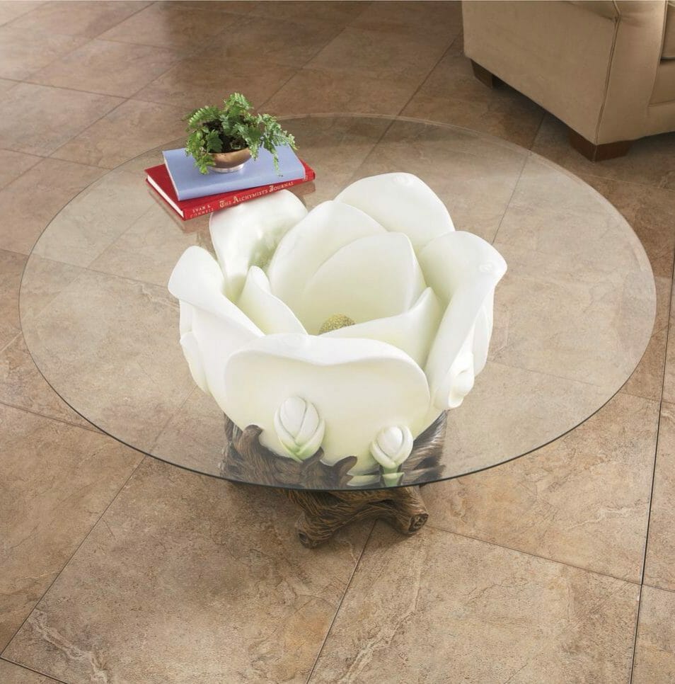 A white blooming rose side table with a round glass top, exposing the flower beneath, with books and a plant on top.