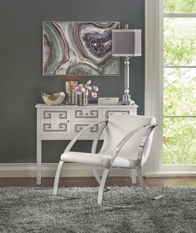 A wall canvas of a purple geode above a white and silver console, a chrome and white chair, and a silver table lamp. 