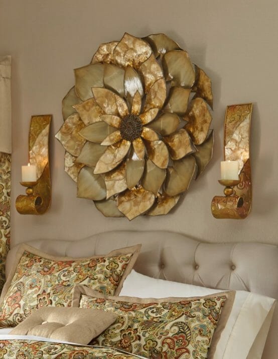 A large capiz wall flower with two matching sconces with lit candles, above a bed with a tufted linen headboard.