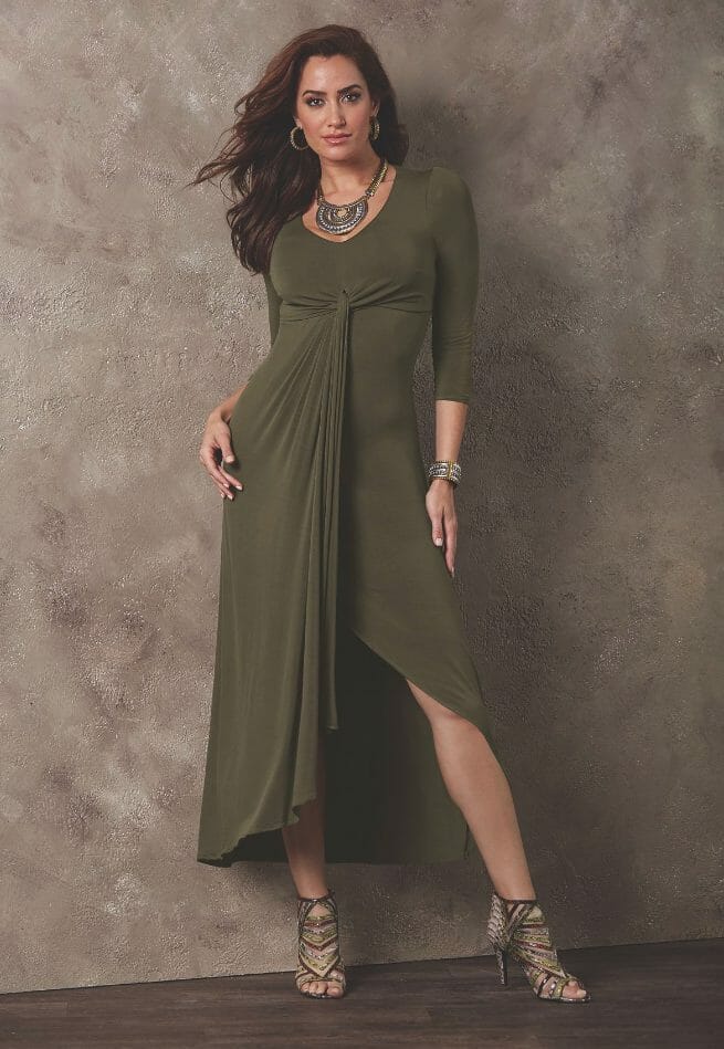 A long-haired woman in a moss green fitted wrap dress with front high-low draping, gunmetal necklace and gladiator shoes.