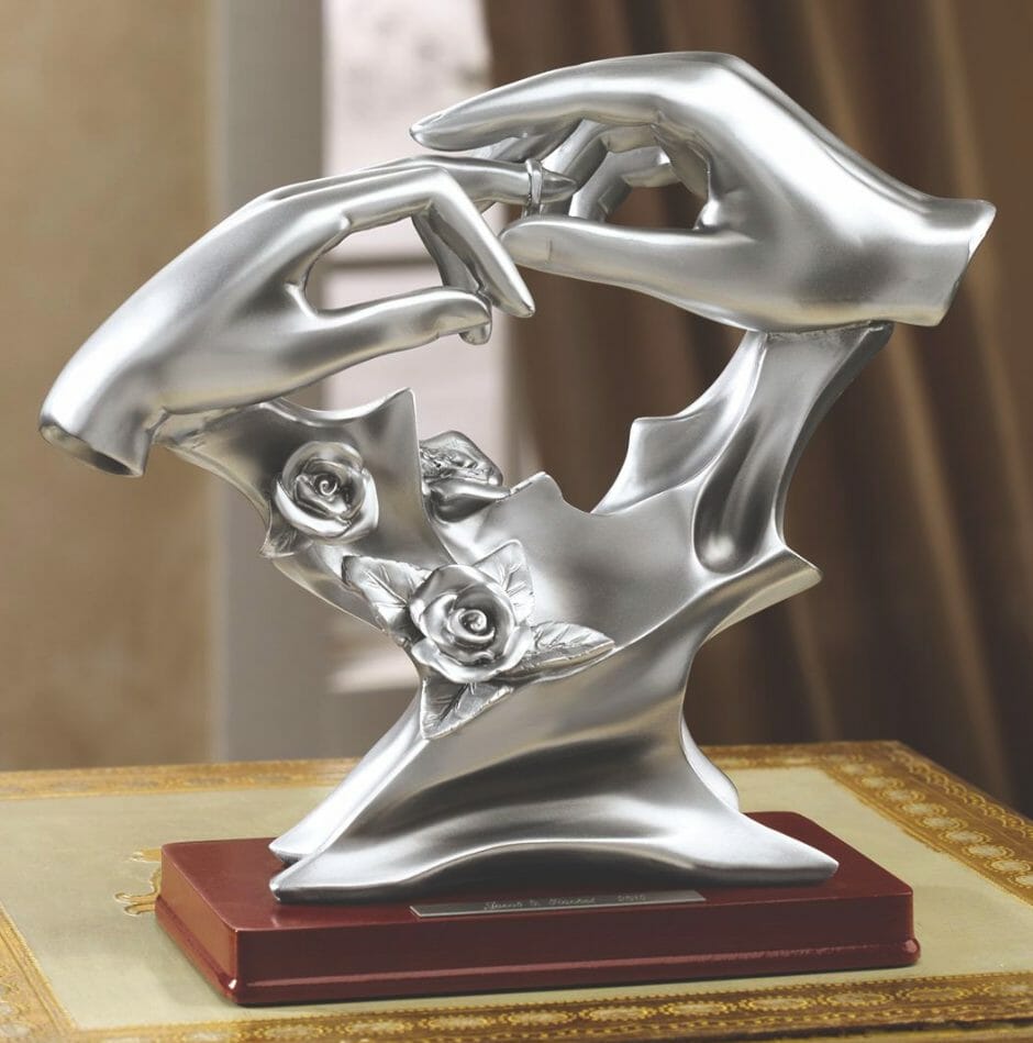 A silver wedding figurine mounted on a wood stand, with two hands, one placing a ring on the other.