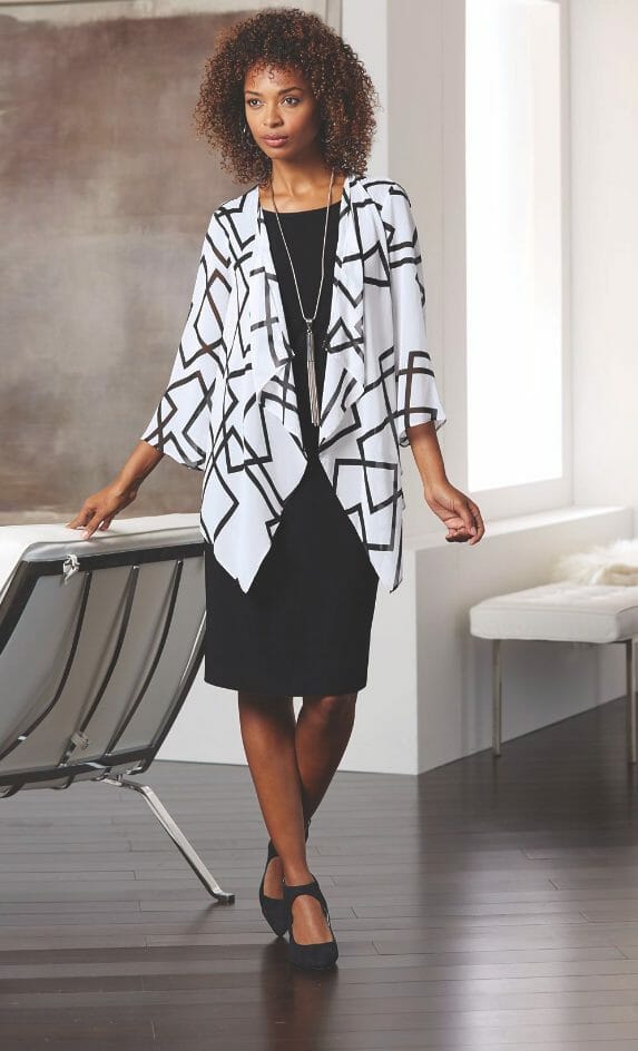 A black woman wearing a black dress with a white and black graphic design loose cardigan and long silver tassel necklace.