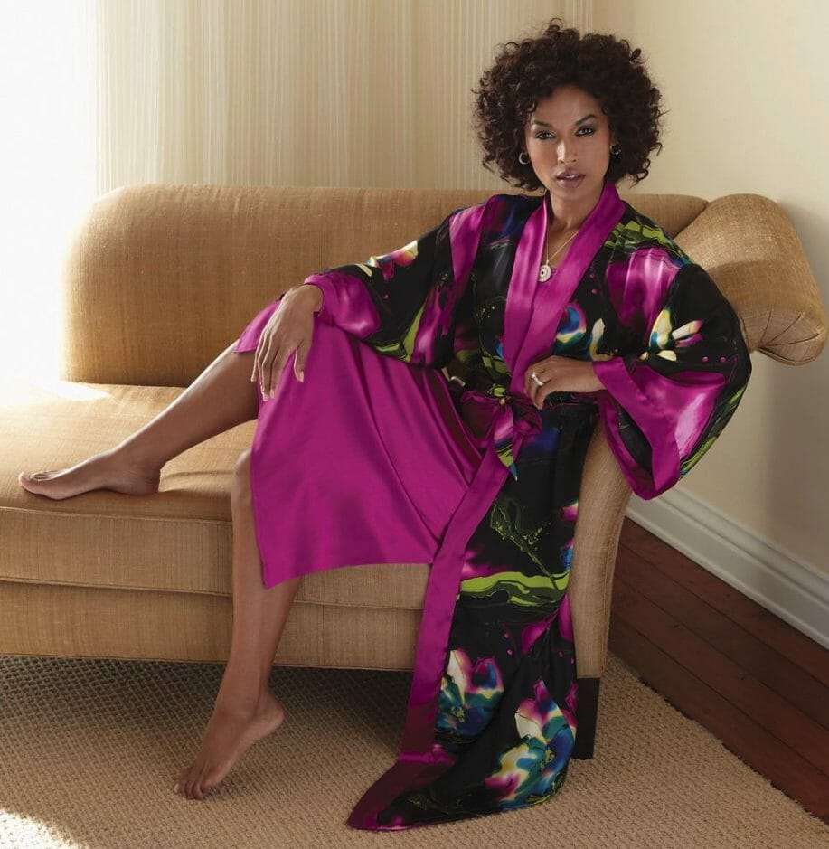 A barefoot African-American woman lounging in a silky black and magenta floral print robe and matching magenta gown.