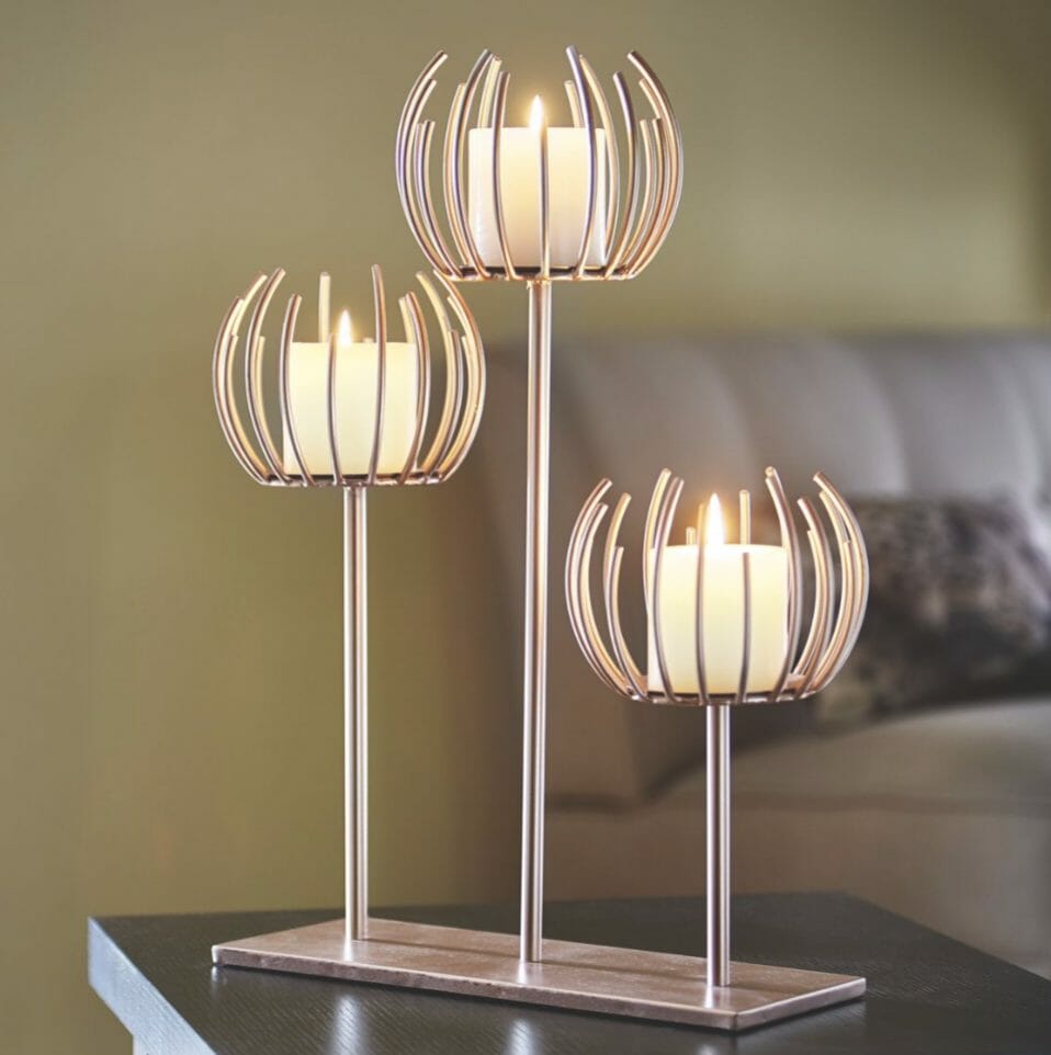 A trio of rose gold metal candle holders mounted on a flat rose gold base, holding three lit votive candles.
