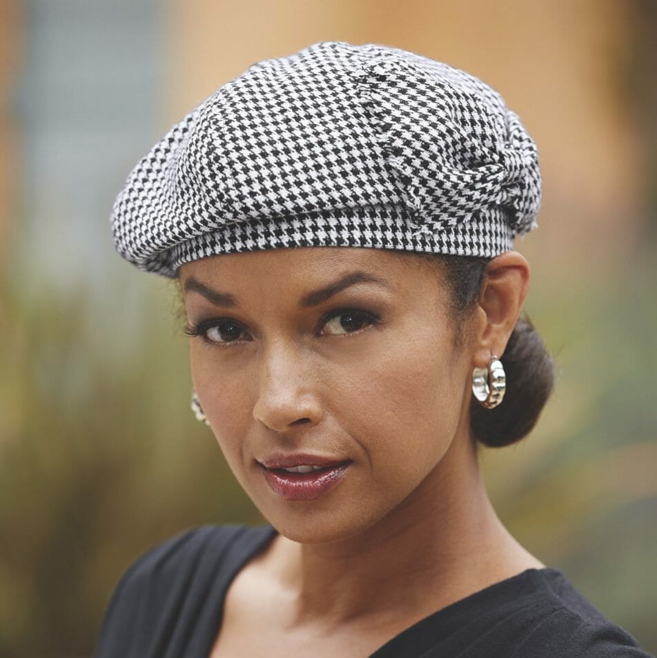 A brown-eyed woman wearing a black and white herringbone newsboy cap, hammered silver earrings, and a black top.