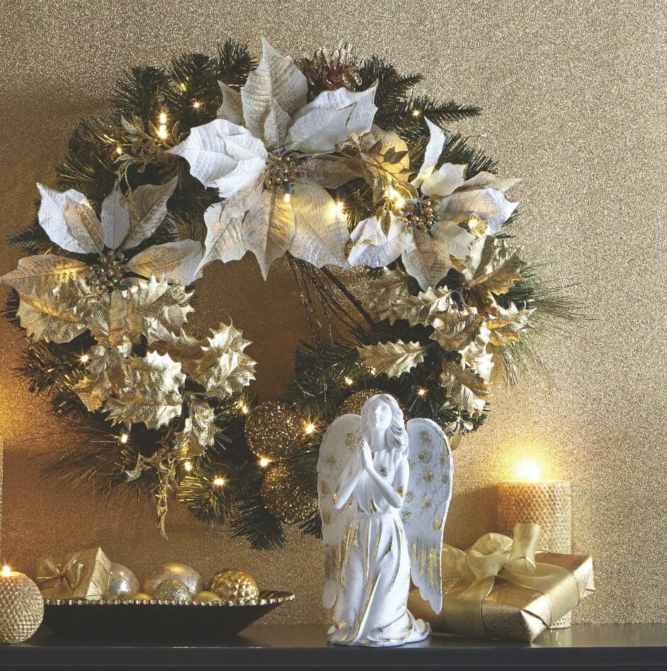 A mantel display of a lit white and gold poinsettia wreath, gold lit candles, wrapped presents, and a white praising angel.