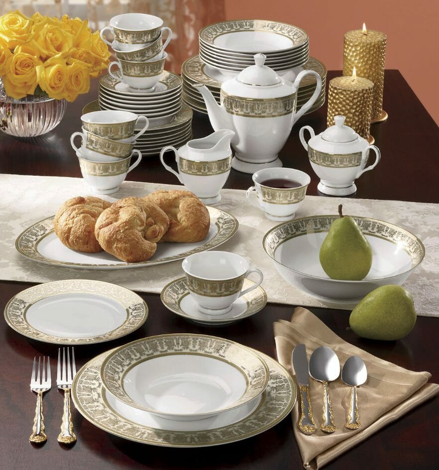 Eight place settings set of gold-rimmed white dinnerware with serving pieces, vased yellow roses, and lit gold candles.