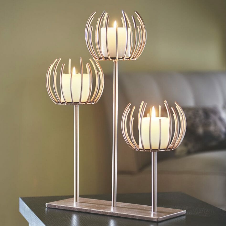A trio of rose gold metal candle holders mounted on a flat rose gold base, holding three lit votive candles.