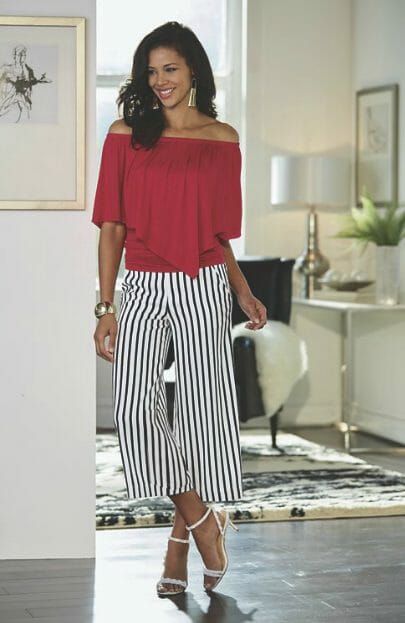 A smiling Hispanic woman wearing a red off-the-shoulder top with a pleated flounce and black and white striped gaucho pant.