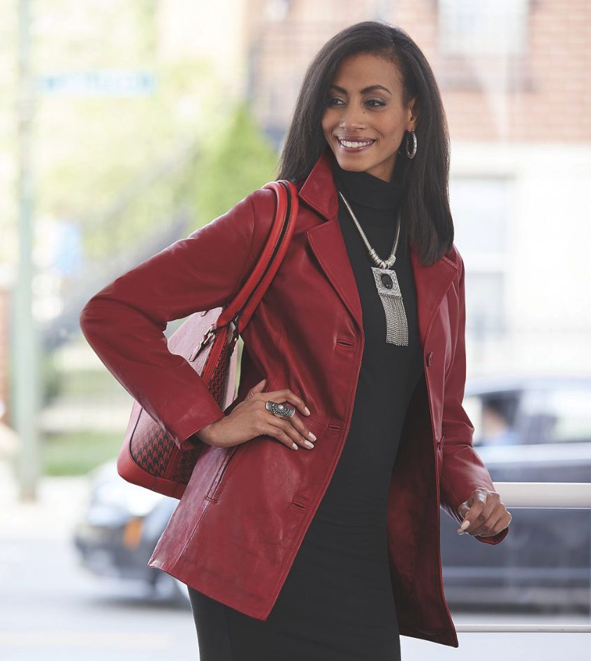 A smiling black woman with a red bag, in a fitted black dress, artistic silver necklace, and a faux leather red jacket.