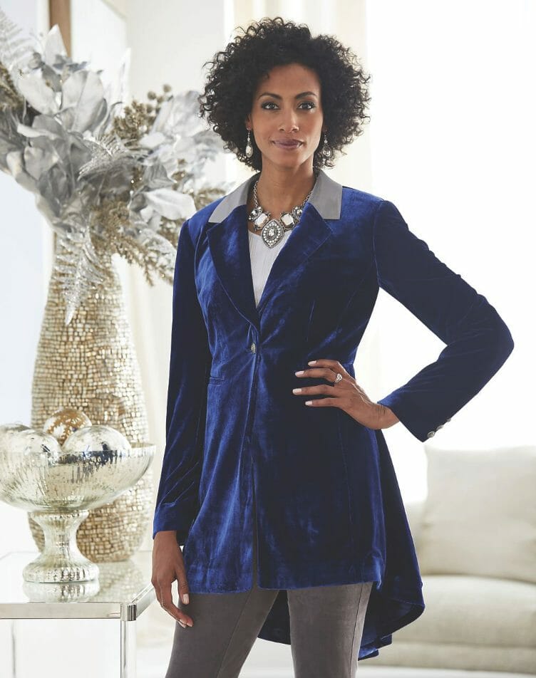 A black woman with curly hair, in a blue velvet high-low jacket, a white top with a designer silver necklace, and gray pant.