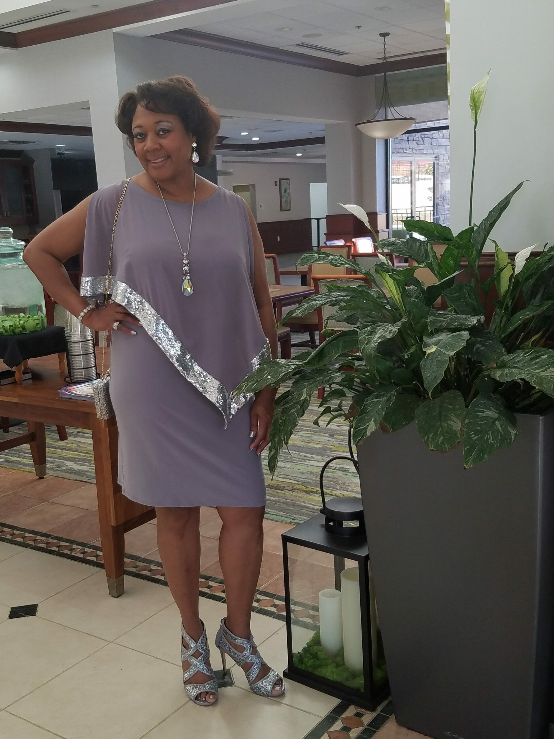 A Midnight Velvet customer at home, in a lavender dress with silver trim on the cape-like top, and silver heeled sandals.