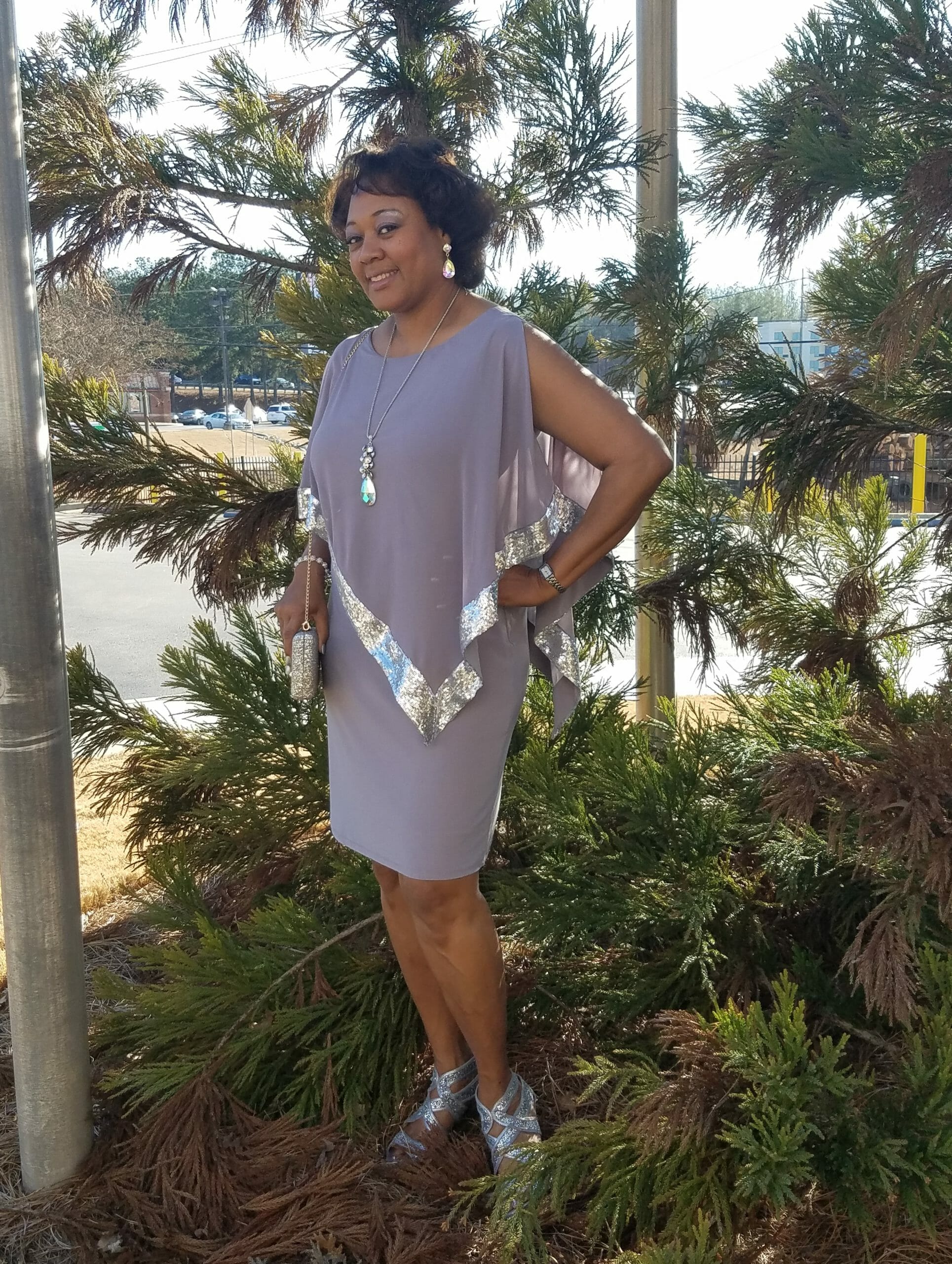 A smiling Midnight Velvet customer by pines, in a lavender dress with silver trim on the cape-like top, and silver sandals.
