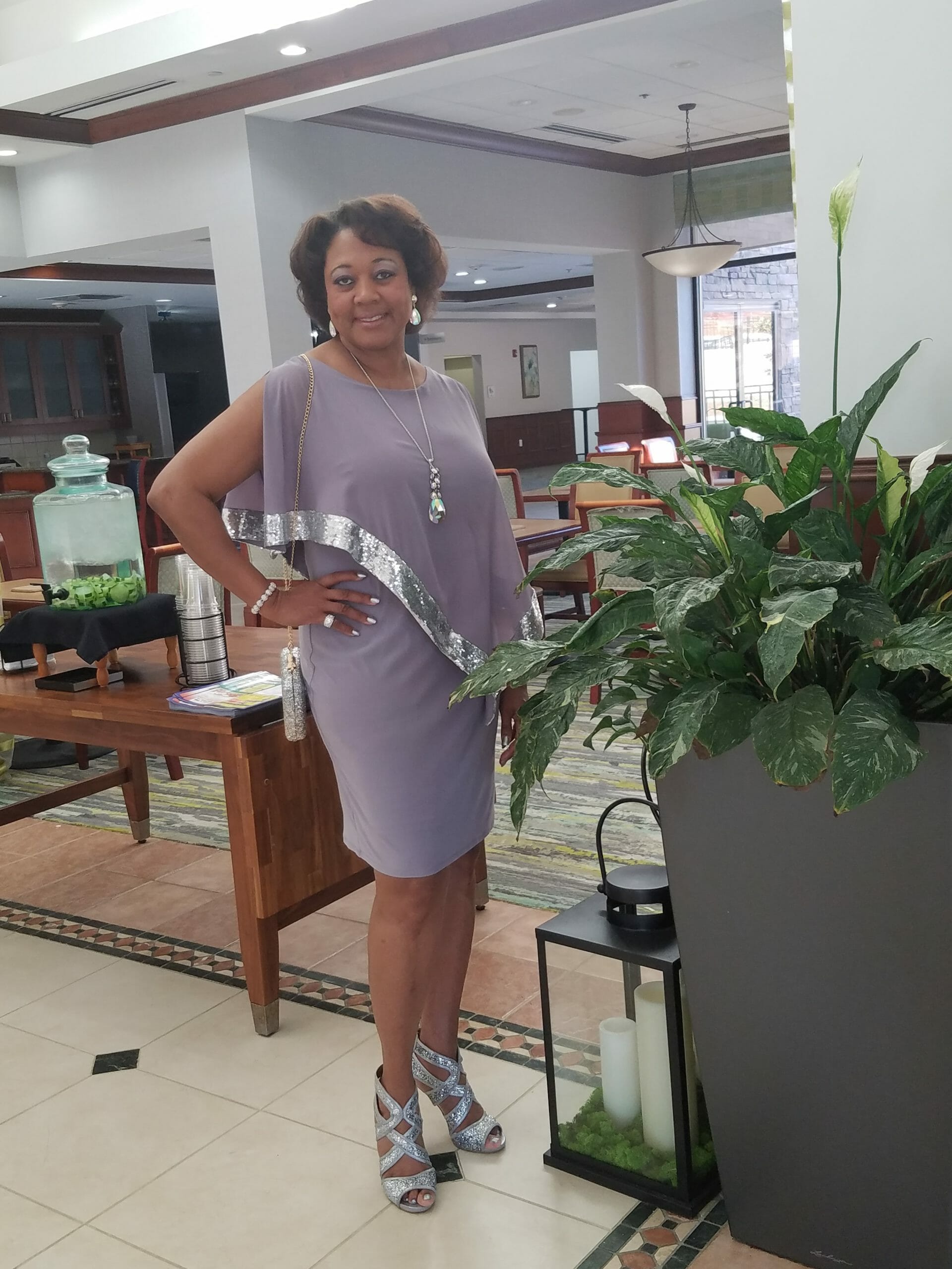 A smiling Midnight Velvet customer, in a lavender dress with silver trim on the cape-like top, and silver heeled sandals.