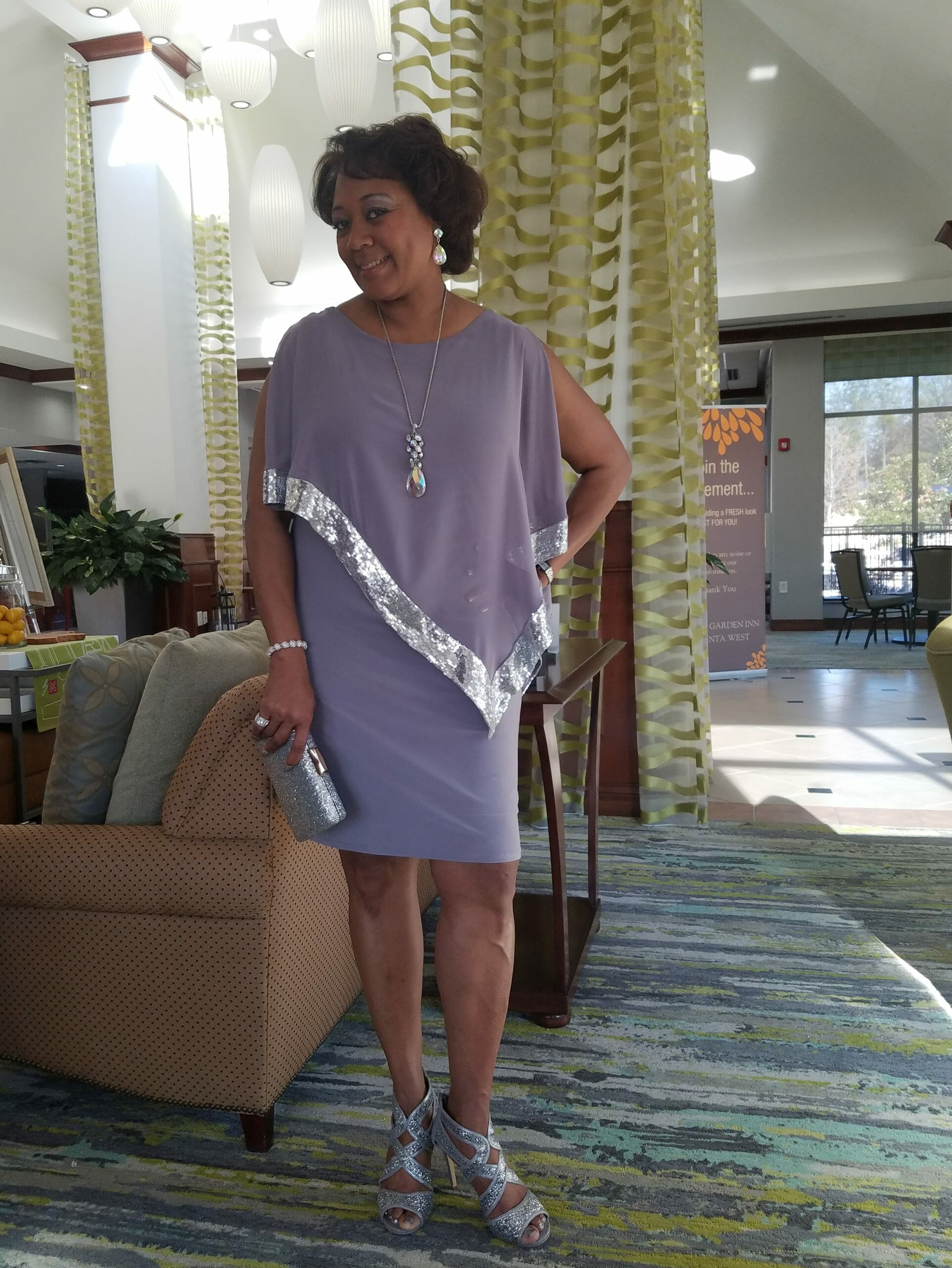 A smiling Midnight Velvet customer, in a lavender dress with silver trim on the cape-like top, and silver heeled sandals.