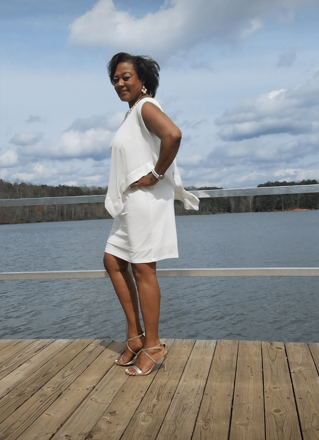 Side view of a smiling Midnight Velvet customer, in a white dress with satin trim on the cape-like top, and silver sandals.