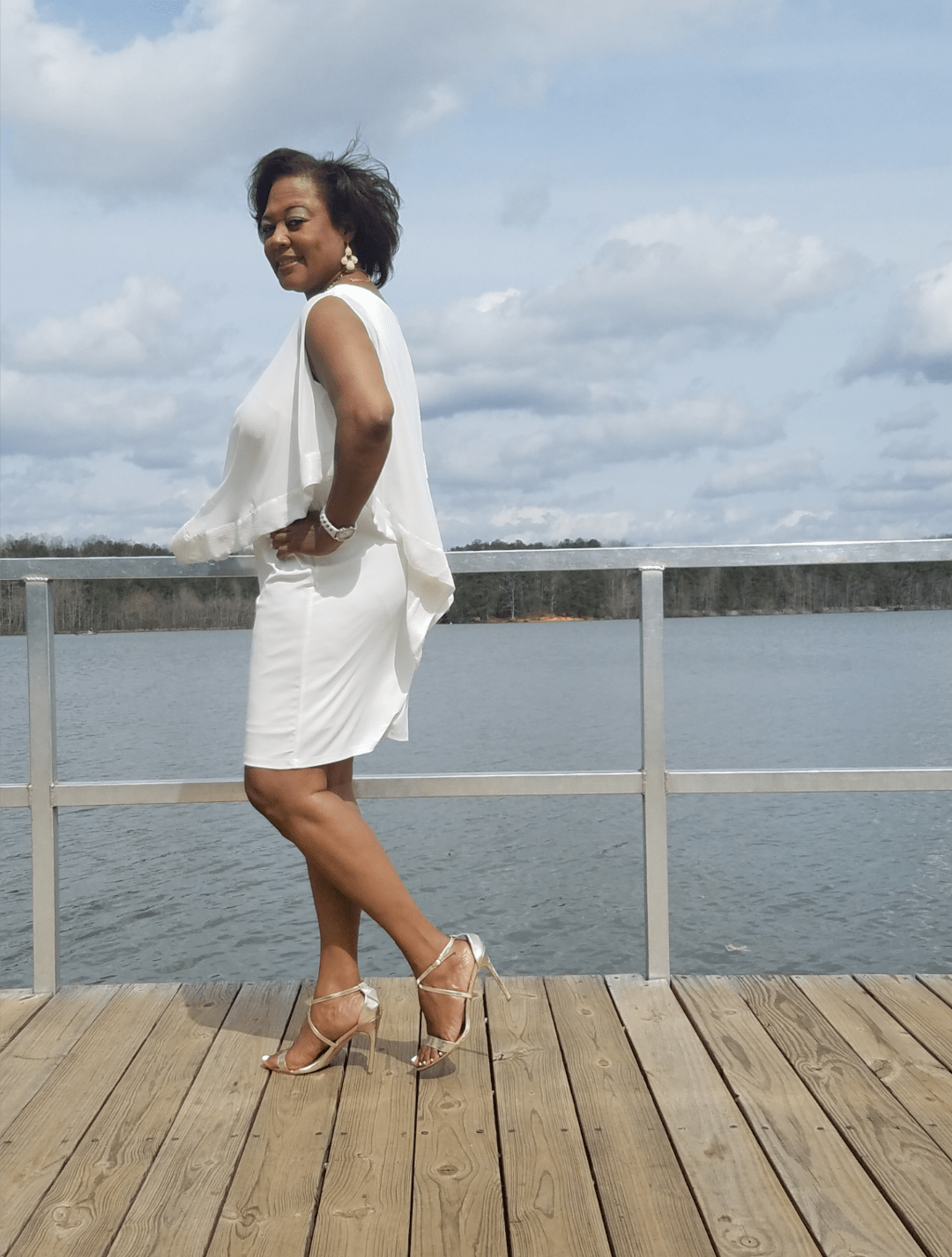 Side view of a smiling Midnight Velvet customer, in a white dress with satin trim on the cape-like top, and silver sandals.