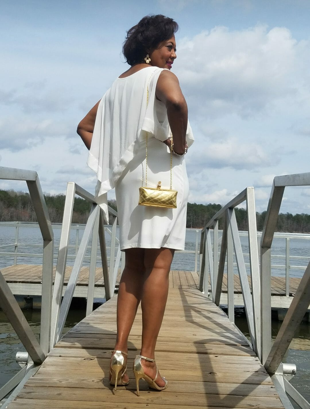 Back view of a Midnight Velvet customer on a pier, in a white dress with satin trim on the cape-like top, with a gold clutch.
