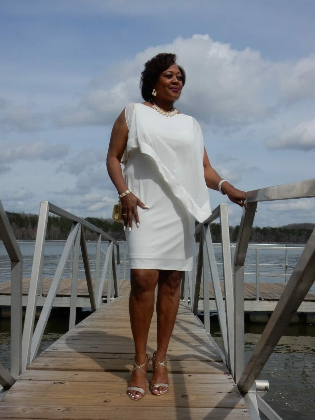 A Midnight Velvet customer on a pier, in a white dress with satin trim on the cape-like top, and silver heeled sandals.
