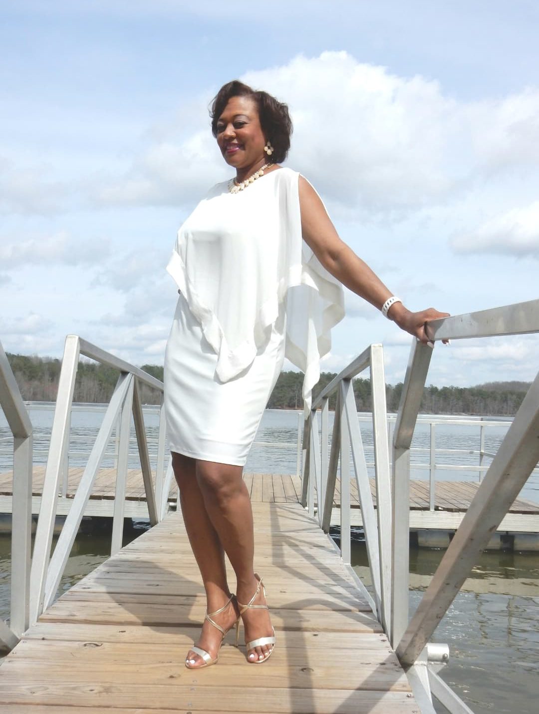 Side view of a Midnight Velvet customer on a pier, in a white dress with satin trim on the cape-like top, and silver sandals.