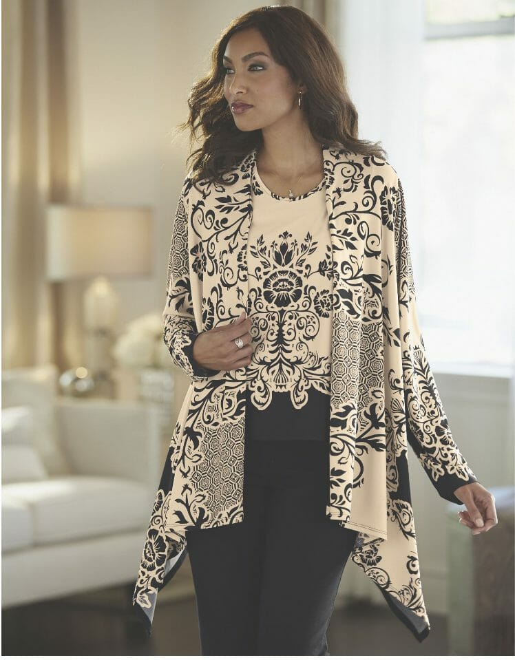 A long-haired black woman in a beige and black floral print top and matching asymmetrical cardigan, and black pant. 