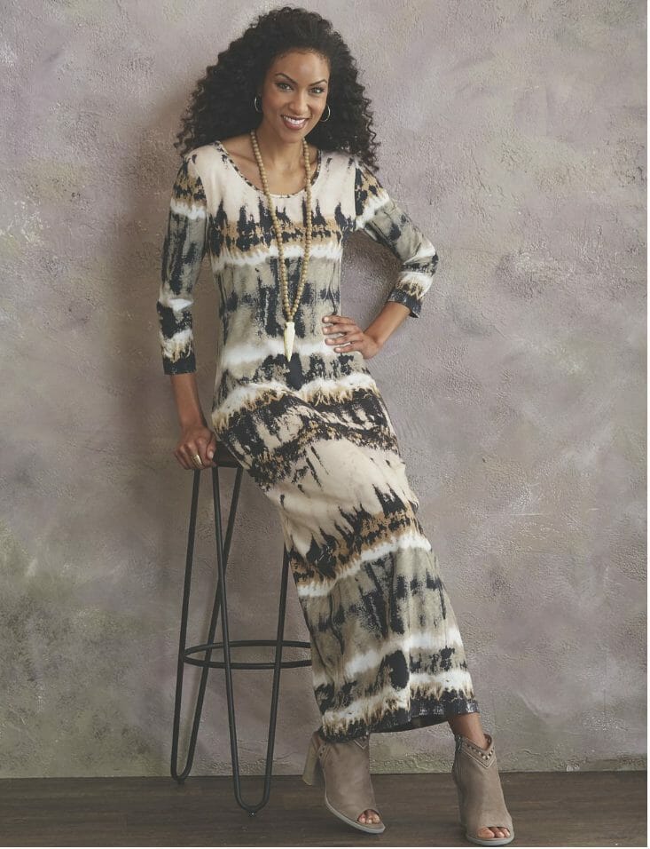 A smiling long-haired black woman in a fitted ivory, beige, gray and black print maxi dress, with faux suede gray booties.