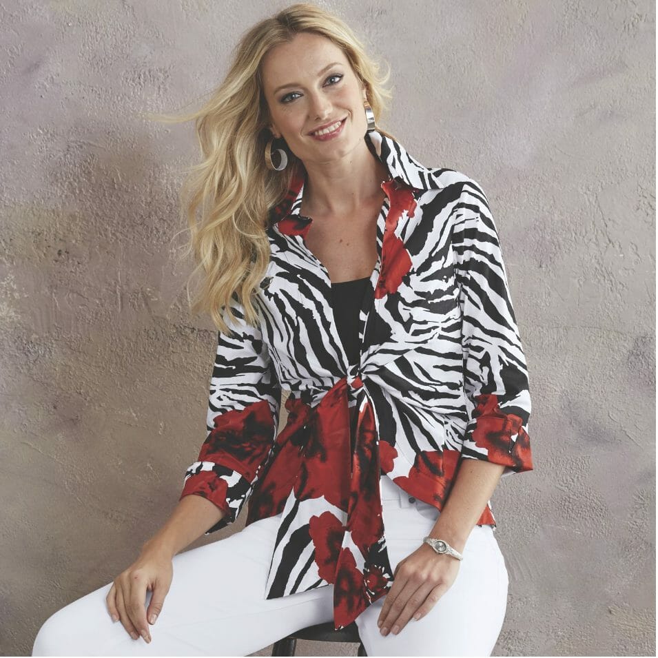 A smiling blonde woman in a zebra stripe and red floral print top with a front tie, a black cami, and white skinny jeans.