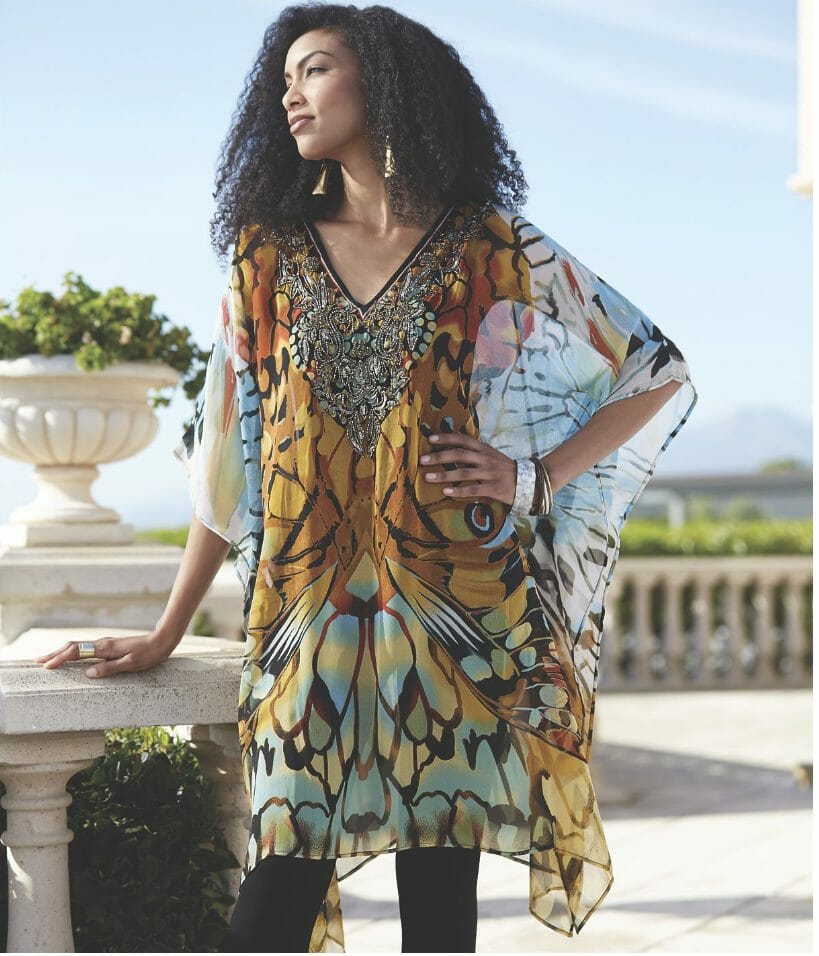 A black woman with long curly hair in a sheer Afrocentric caftan top with beaded neckline, and black leggings. 