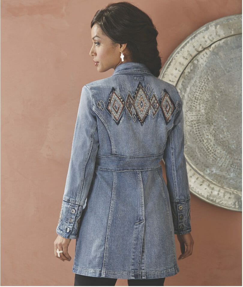 Back view of a brunette woman in a washed denim coat with baroque patterned diamond shapes embroidered on the back.