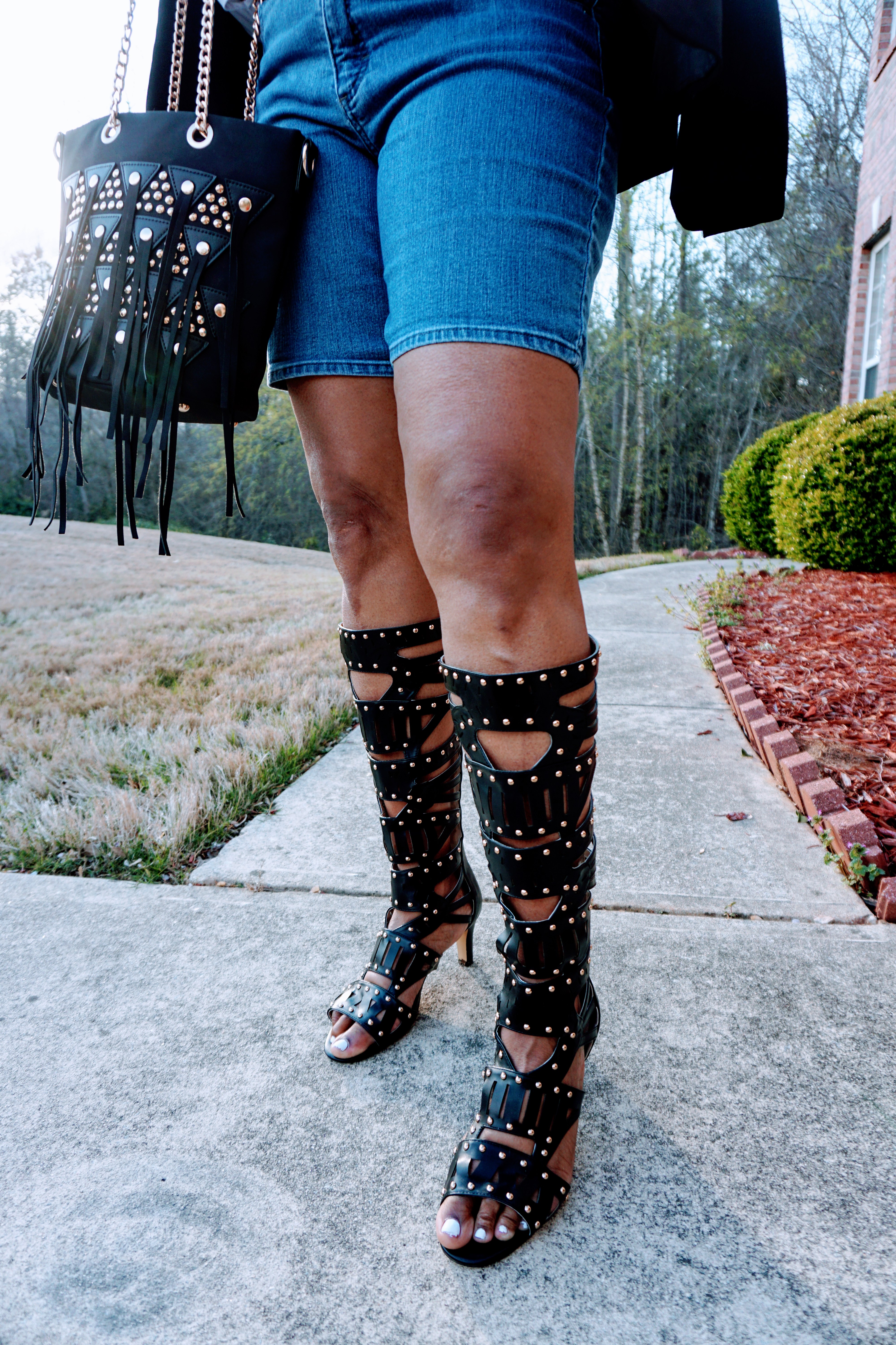 Lower half of a woman in tall black studded boots, and carrying a matching black studded and fringed bag.