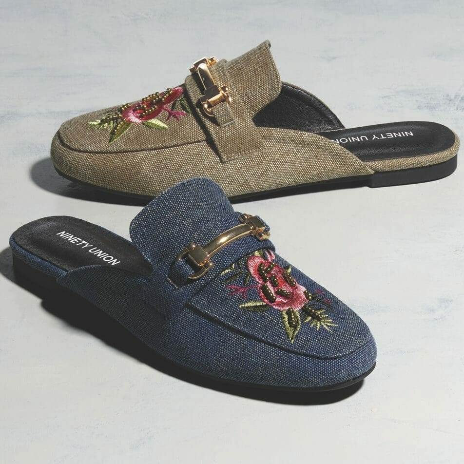 Two canvas mule shoes with rose embroidery and gold buckle detail on the toe, one in denim blue and one in tan.