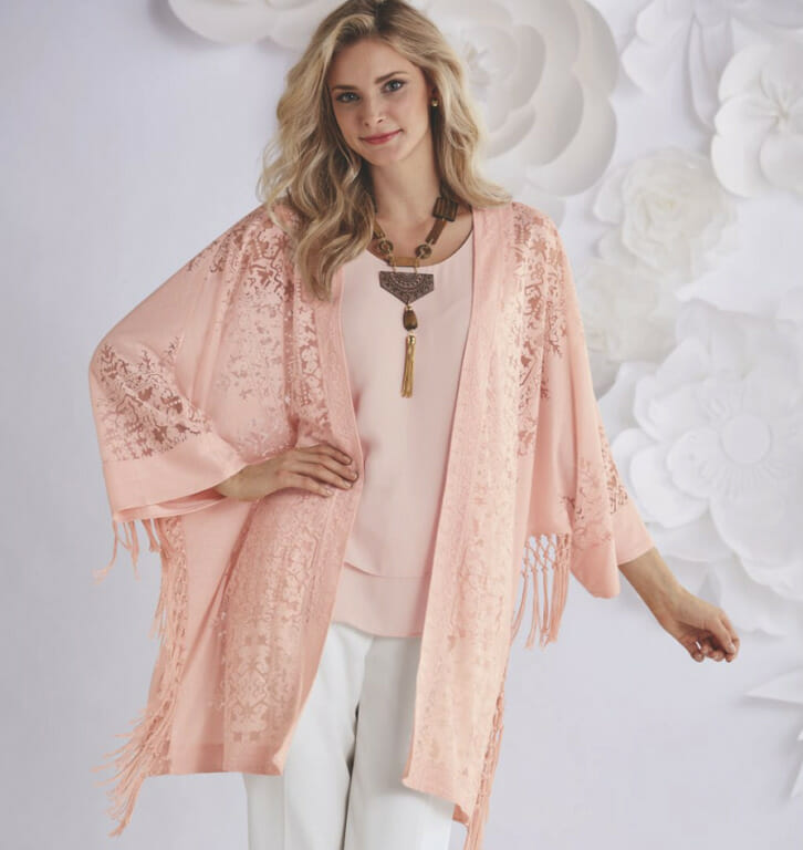 A smiling blonde woman in a dusty rose cami and matching fringed lace caftan-like cardigan, bronze necklace, and white pant.