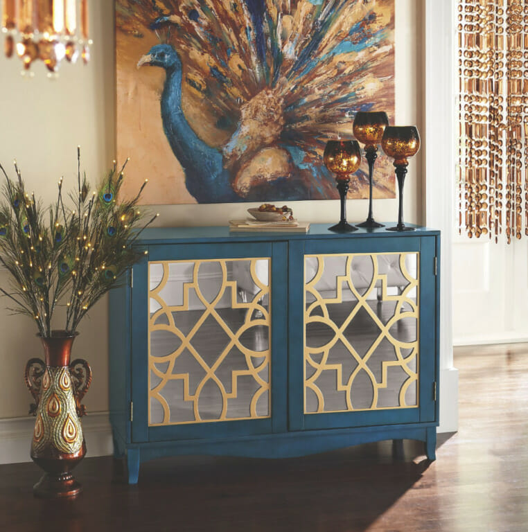 A blue mirrored 2-door credenza with a large peacock canvas in blue and tan, peacock feathers in an urn, and lit candles.
