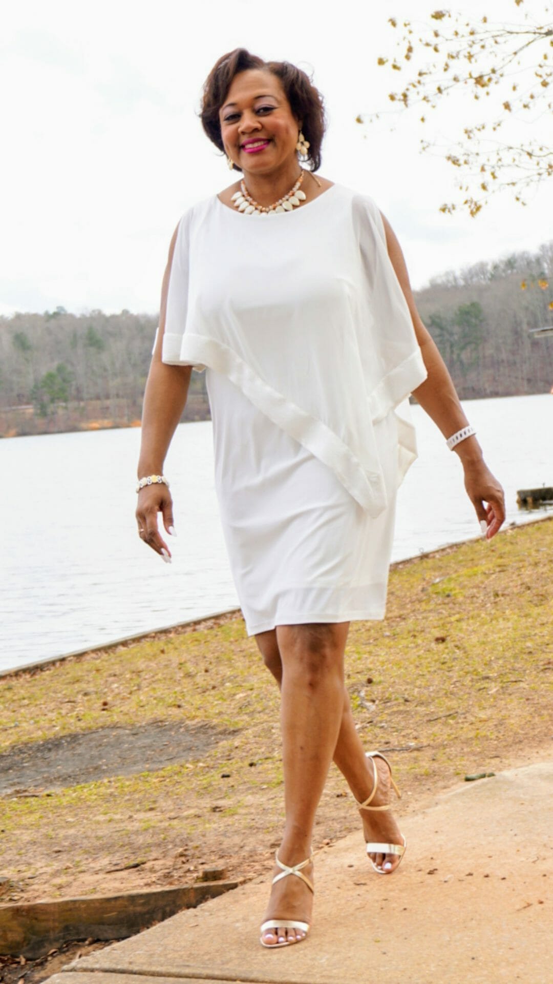 A smiling Midnight Velvet customer lakeside, in a white dress with satin trim on the cape-like top, and silver sandals.