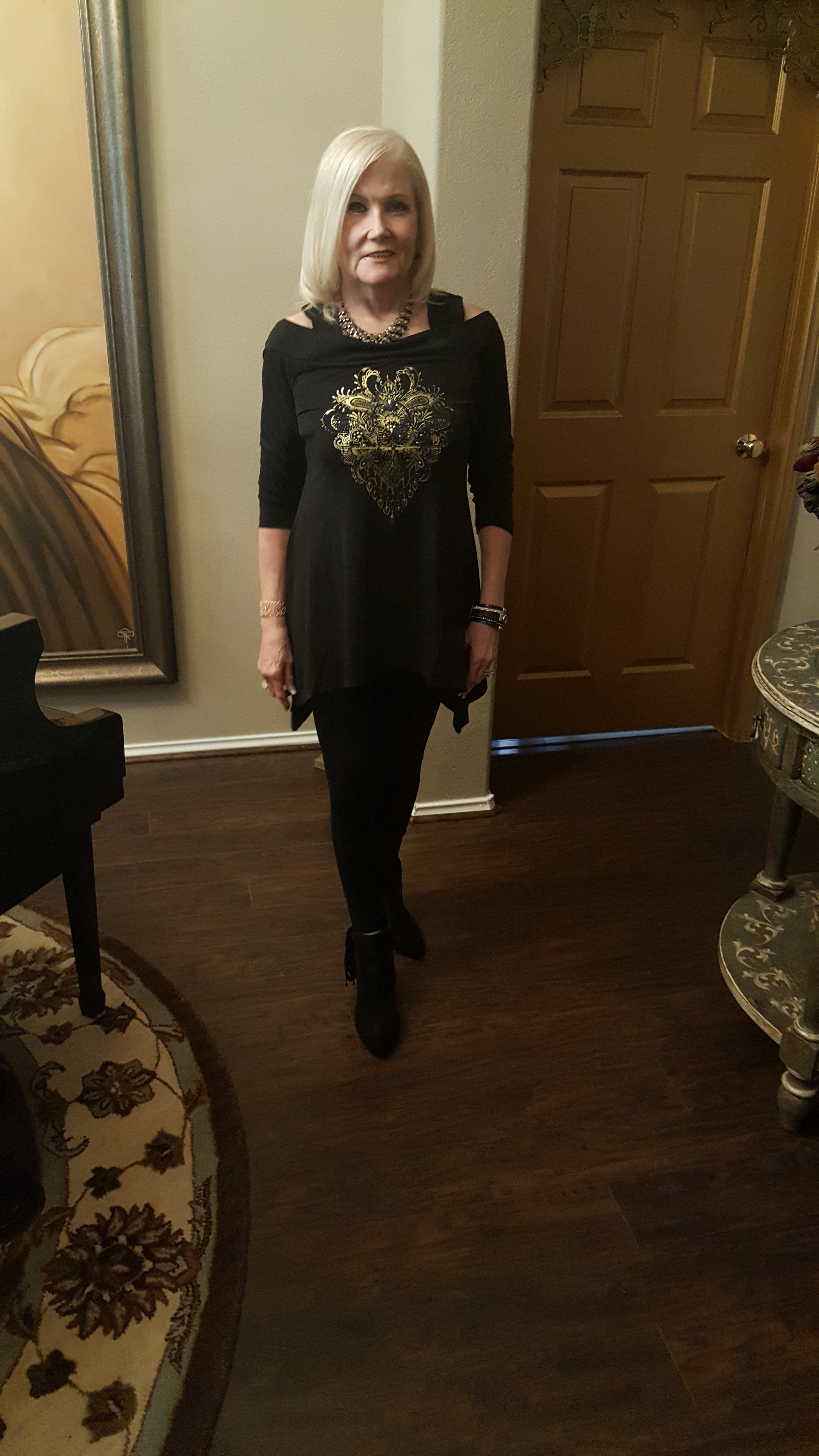 A Midnight Velvet customer in a black cold-shoulder top with a gold floral design, black leggings, and black booties.