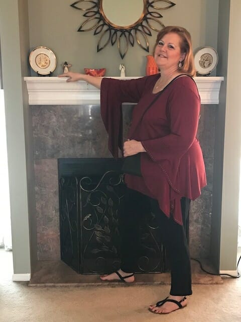 A Midnight Velvet customer by a fireplace, in a burgundy tunic with flowing sleeves, black pant, and flat black sandals.
