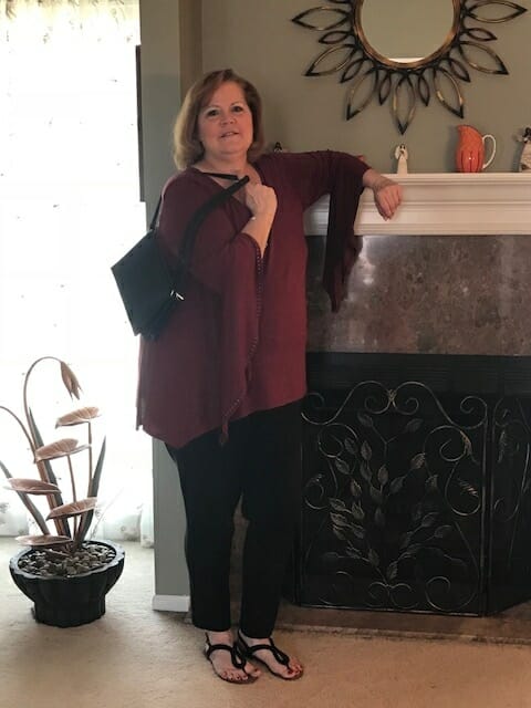 A Midnight Velvet customer by a fireplace, in a burgundy tunic with flowing sleeves, black pant, and holding a black bag.