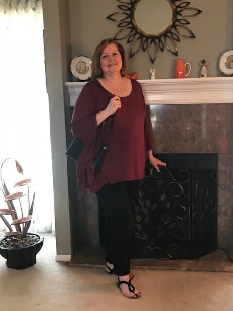 A Midnight Velvet customer by a fireplace, in a burgundy tunic with flowing sleeves, black pant, and holding a black bag.